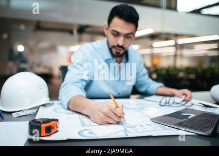 Concentrated serious attractive millennial arab man engineer with beard works with drawings at workplace Stock Photo