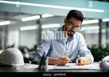 Serious attractive millennial arabic man engineer in glasses with beard works with drawings at workplace with hard hat Stock Photo