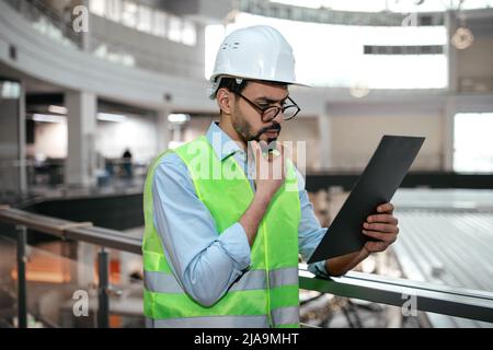 Pensive millennial muslim guy engineer in protective uniform and hard hat in glasses with beard looks at tablet Stock Photo
