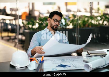 Serious handsome young muslim engineer in glasses with beard with project drawings at workplace with hard hat Stock Photo