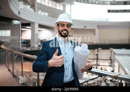 Glad confident millennial islamic guy engineer in protective helmet, suit with beard with project drawings Stock Photo