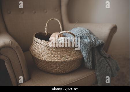 Making homemade knitted sweater at home. Straw basket with yarn wool skein with knitted fabric on armchair indoors. Having hobby. Cozy hygge Stock Photo