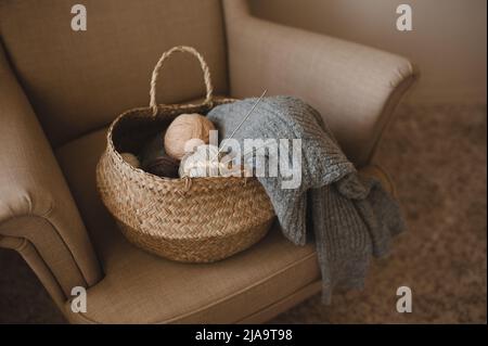 Making homemade knitted sweater at home. Straw basket with yarn wool skein with knitted fabric on armchair indoors. Having hobby. Cozy hygge Stock Photo