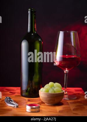 Helsinki / Finland - MAY 29, 2022: An empty red wine bottle, a glass of wine, and some grapes on a wooden surface against a dark background. Stock Photo