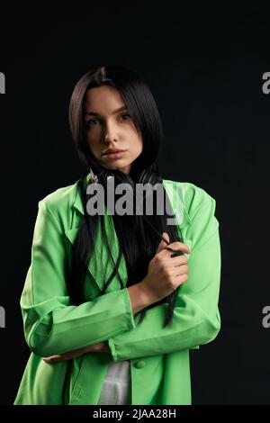 Elegant pretty girl looking at camera with sad expression inside. Portrait view of thoughtful woman in green blazer with headphones on neck, isolated on black studio background. Concept of emotions. Stock Photo