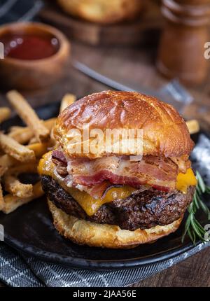 Bacon cheeseburger on a toasted bun and french fries on a black plate Stock Photo