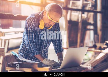 professional carpenter using laptop computer aid design(CAD) technology with wooden furniture handcraft working in wood workshop Stock Photo