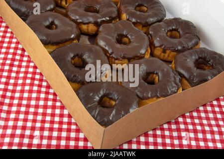 Open box of donuts with chocolate icing on red and white check tablecloth Stock Photo