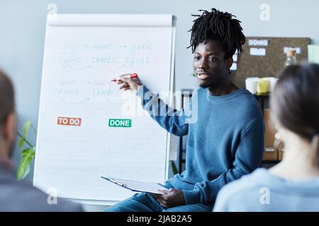 Portrait of young black man teaching English class in office to group of people and pointing at whiteboard with grammar rules Stock Photo