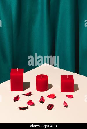 Romantic composition with red candles and red rose petals on bright beige table. Silk green curtain background. Valentines Day concept. Copy space. Stock Photo