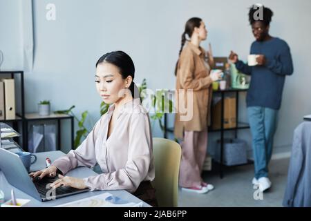 Portrait of young Asian woman using laptop at workplace with people chatting in background in company office, copy space Stock Photo