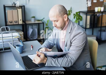 Portrait of balding adult man reading data document and using laptop while working in company office Stock Photo