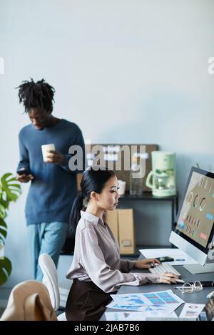 Vertical portrait of diverse team working and operating in company office, focus on Asian young woman using computer Stock Photo