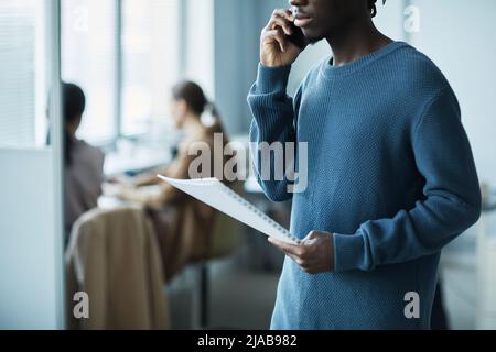 Cropped shot in blue of young African American man speaking on phone in office setting and holding documents, copy space Stock Photo