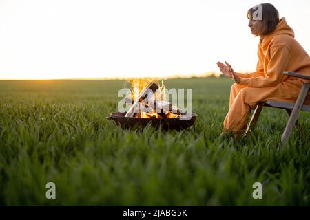 Woman sitting by the fireplace on green field on sunset Stock Photo