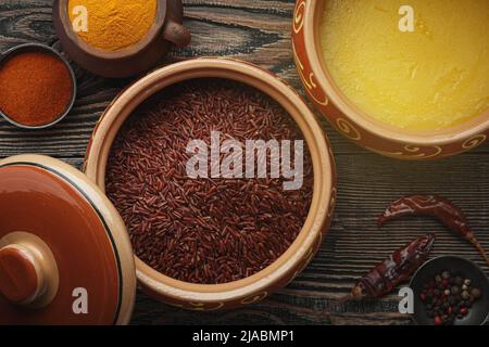 Ghee or clarified butter in ceramic bowls and red rice with different spices on an old wooden table. Top view. Stock Photo