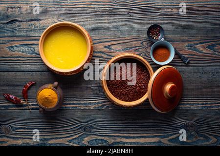 Ghee or clarified butter in ceramic bowls and red rice with different spices on an old wooden table. Top view. Stock Photo