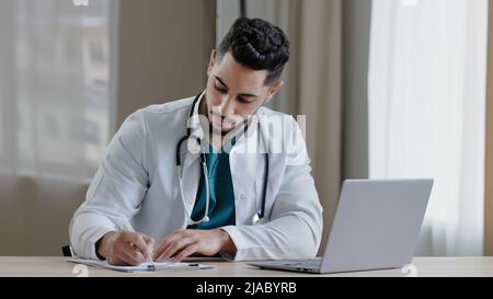 Concentrated arabian man doctor practitioner sit at clinic cabinet use computer chatting distant in medical social app give online consultation Stock Photo