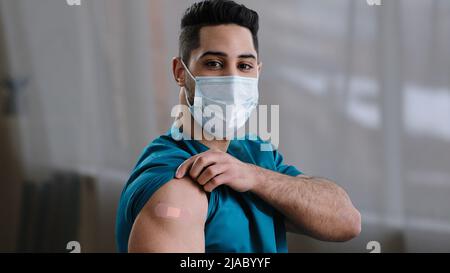 Inspired arabian hispanic man young male practitioner nurse intern in medical face mask showing injection mark adhesive bandage shoulder make vaccine Stock Photo