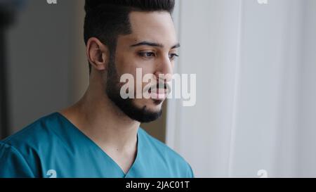 Young pensive hispanic doctor male thoughtful arabian nurse practitioner therapist surgeon stand near window in hospital workplace wait for patient Stock Photo