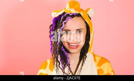 Young cute woman with dreadlocks in kigurumi looking at camera and showing tongue. Funny playful female with curly hairstyle in pajamas on pink backgr Stock Photo