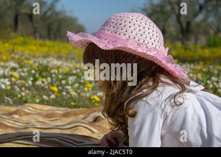Image of an adorable little girl in a pink hat lying on a blanket in the middle of a field of flowers during a vacation. Stock Photo