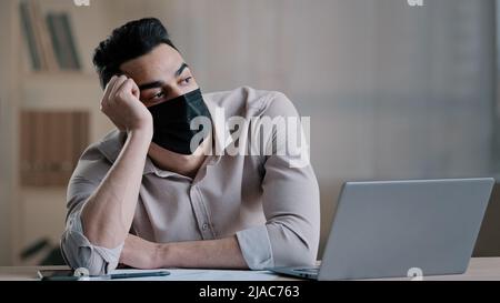 Bored sad hispanic worker young business man in medical mask tired unmotivated employee disinterested in dull distant work on computer lazy male arab Stock Photo