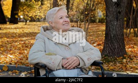 Happy elderly granny sit in wheelchair looks up in autumn park adult retired woman resting outdoors person with disability smiling enjoying good Stock Photo