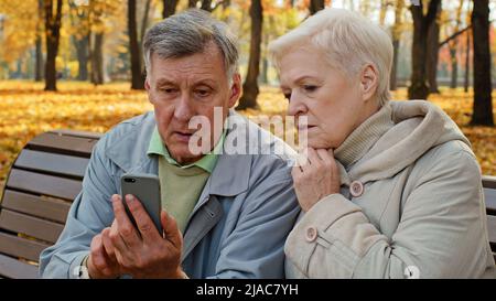 Upset old married caucasian couple use smartphone sit on bench in autumn park look at screen phone worried elderly retired people sad family feel Stock Photo