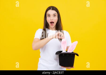 Happy Easter. Portrait of young excited woman with bunny ears isolated on yellow studio background. Rabbit ears appear from the magic hat. Stock Photo