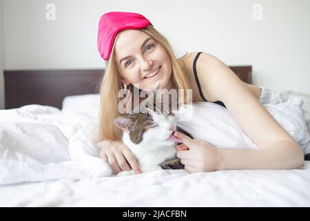 Young blonde 30s woman with sleep mask and cute cat lying on bed in the room Stock Photo