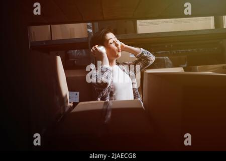 Silly playful female warehouse worker using cups as headphones through shelves with boxes, looking at the camera. Stock Photo