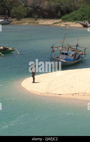 A fisherman standing on sandy beach in a bacground of fishing boats on a lagoon-like seascape in the fishing beach of Pero in Pero Batang villlage, Kodi, Southwest Sumba, East Nusa Tenggara, Indonesia. Stock Photo