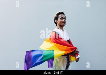 Side view of smiling male with closed eyes holding LGBT rainbow flag against grey wall Stock Photo