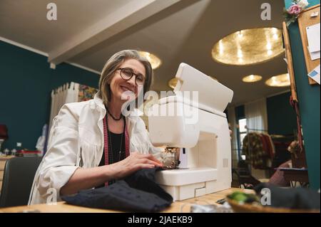 Mature female dressmaker, seamstress, tailor or sewer in workshop studio, works on sewing machine, designing new collection clothes. Fashion designer Stock Photo