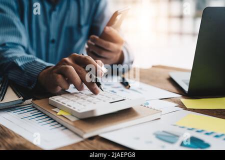 Close up of businessman or accountant hand holding mobile phone working on calculator to calculate business data, accountancy document and laptop Stock Photo