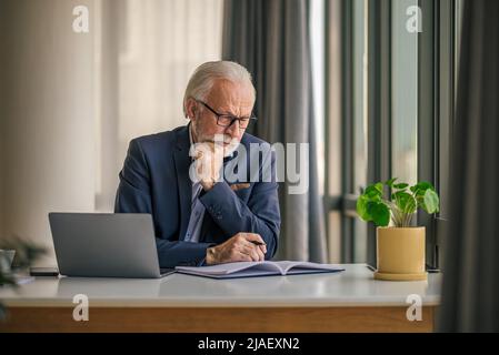 Confident senior businessman making notes, thinking and planning. Male professional is working on laptop at desk he is multitasking while sitting at o Stock Photo