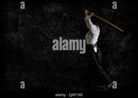A girl in black hakama standing in fighting pose with wooden sword bokken over black background. Shallow depth of field. Grunge. Stock Photo