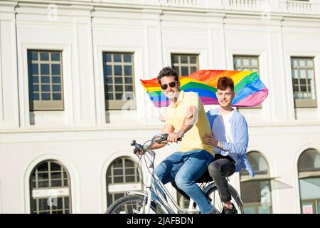 Young gay men couple riding bicycle with gay pride flag looking at camera outdoors. lgbt concept Stock Photo