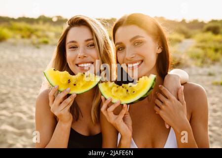 Young two women eating watermelon and hugging during picnic on beach Stock Photo