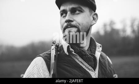 Portrait of hunter man in traditional shooting clothes on field holding shotgun, black and white photo. Stock Photo