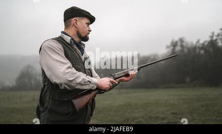 Hunter man in traditional shooting clothes on field aiming with shotgun. Stock Photo