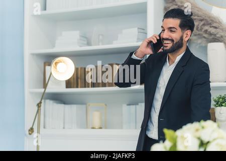 Young spontaneous handsome bearded Cuban entrepreneur man in a black suit in an office talking via his modern smartphone and smiling. High quality photo Stock Photo