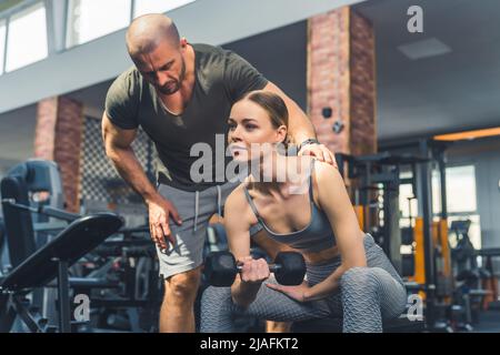 Sportive European woman doing shoulder exercises with dumbbell on a bench at gym while her personal coach corrects her posture. High quality photo Stock Photo