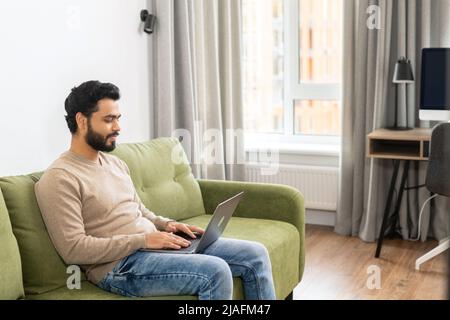 Positive Hindi man in smart casual clothes using computer while sitting at the sofa in his flat. Young Indian male student watching webinars, educational courses, learning on the distance Stock Photo