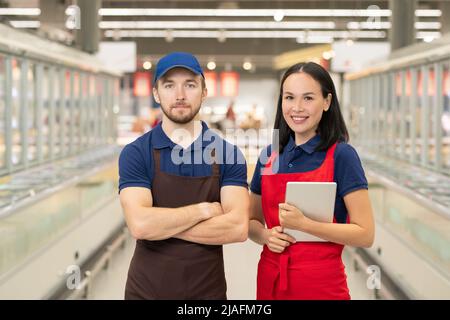 Horizontal medium portrait if man and woman wearing uniform standing together in supermarket aisle looking at camera Stock Photo