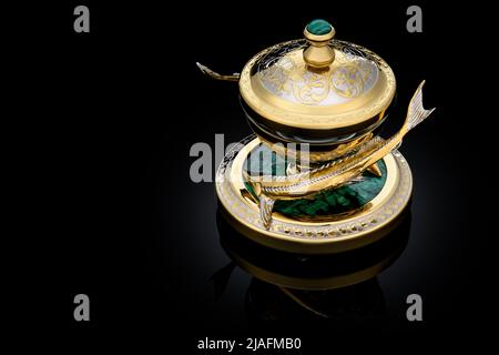 Gilded luxury crystal caviar bowl on stand made of natural quartz, carved brass fish figurine on a black background. Stock Photo