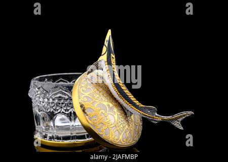 Gilded luxury crystal caviar bowl on stand made of natural quartz, carved brass fish figurine on a black background. Stock Photo