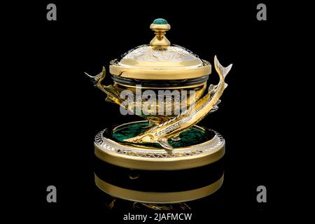 Gilded luxury crystal caviar bowl on stand made of natural quartz, carved brass fish figurine on a black background. Stock Photo