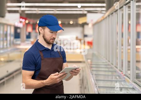 Young man wearing uniform working as merchandiser in modern supermarket using digital tablet, horizontal medium shot Stock Photo